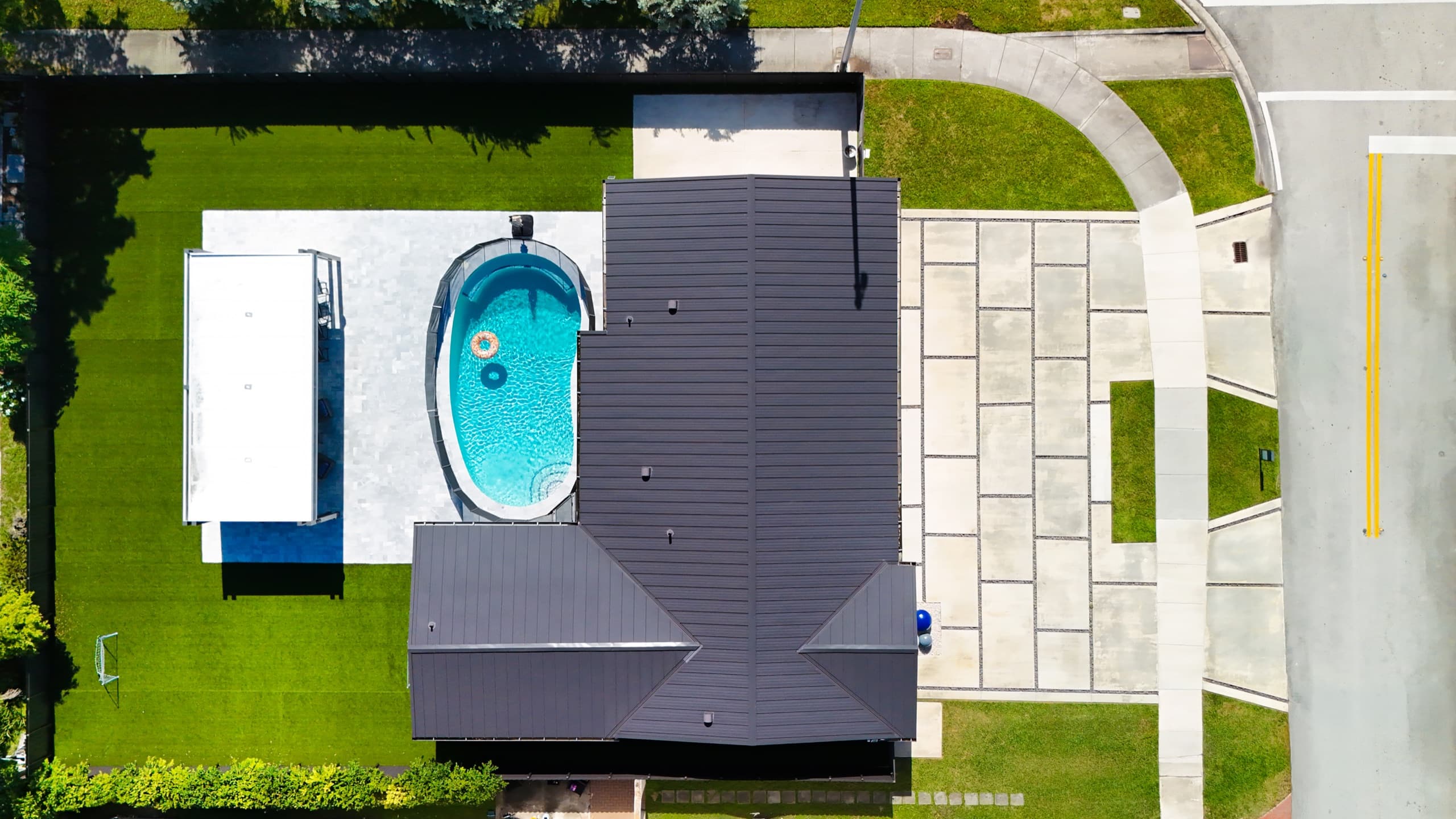 Top-down aerial view of the house: a dark roof, a turquoise pool and a white-roofed pavilion in the backyard, and a driveway of large concrete slabs.