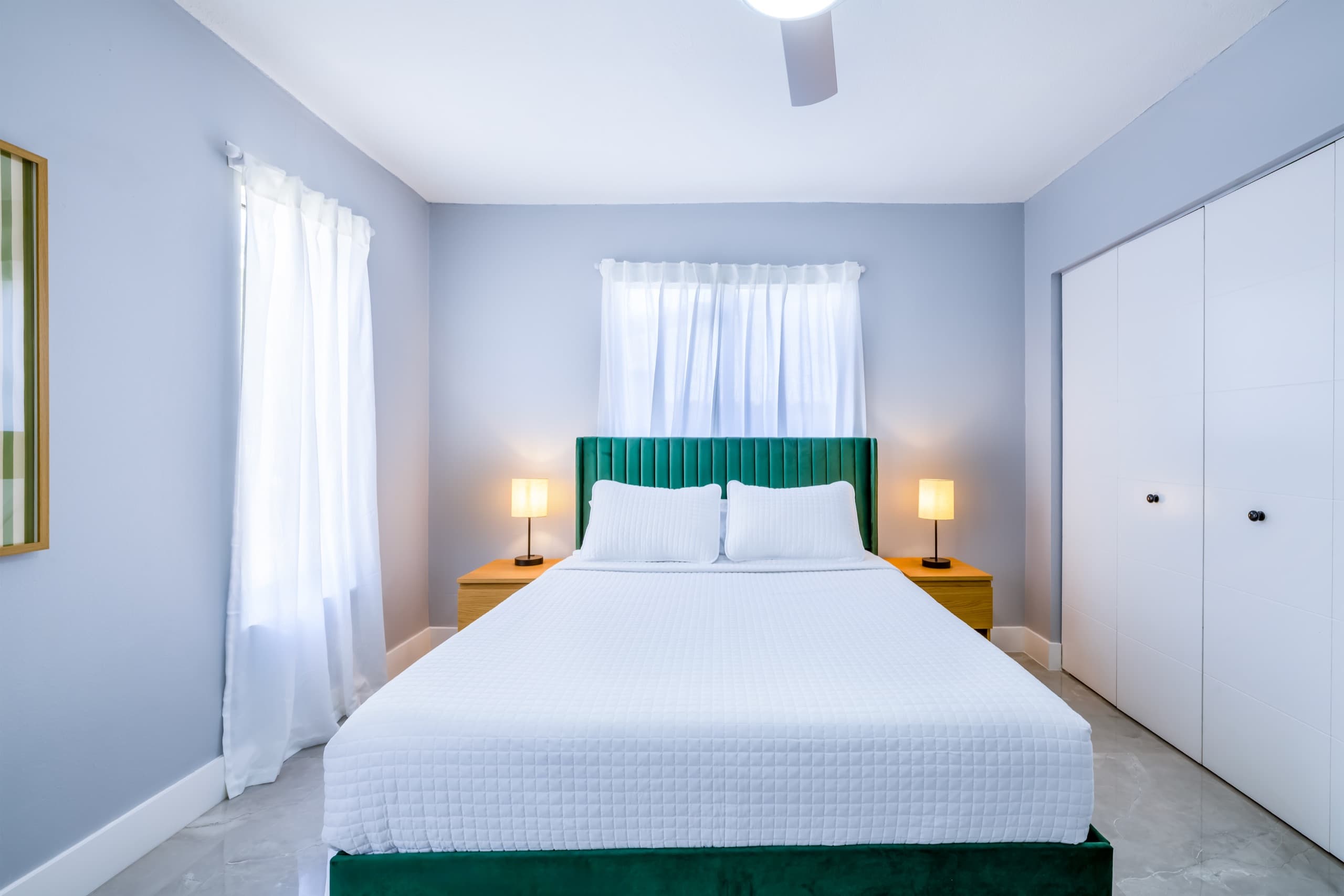 Queen bed with an emerald upholstered headboard between two oak nightstands with lit lamps, under a window with white curtains.
