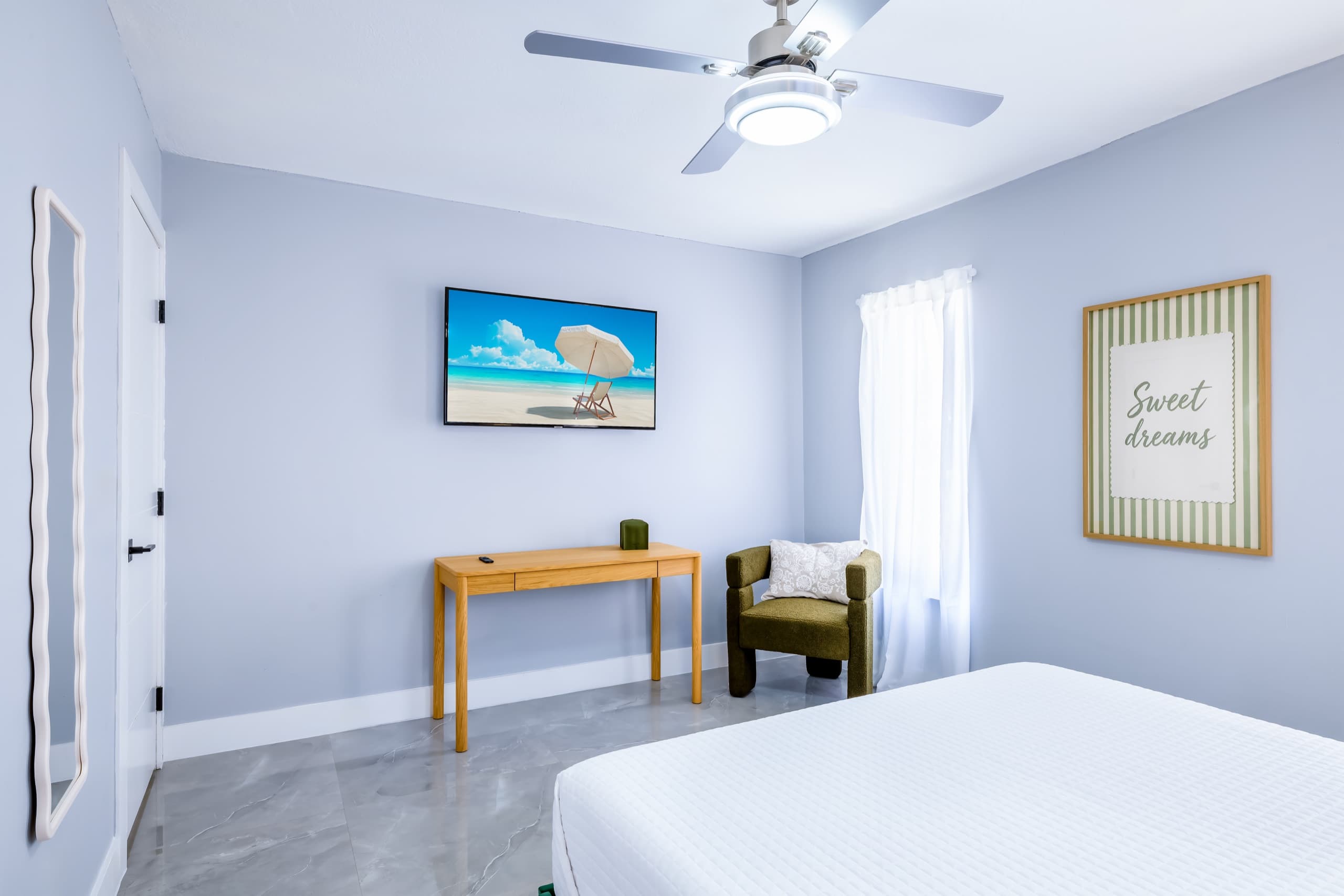 Desk, olive armchair and wall-mounted TV opposite the bed, beside the bedroom door and a wavy full-length mirror.