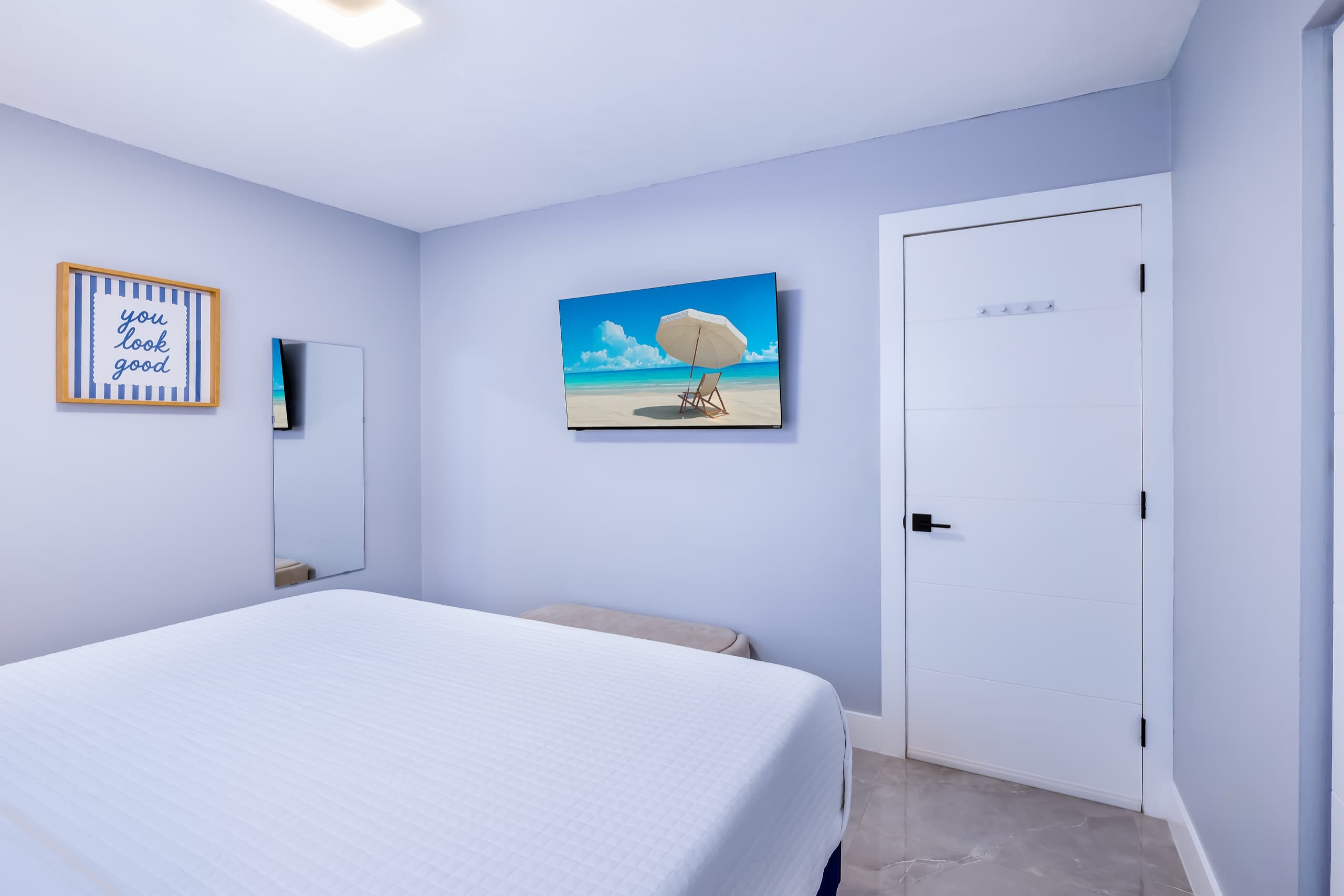 Wall-mounted TV and bedroom door seen from beside the bed, with a full-length mirror and a framed print.