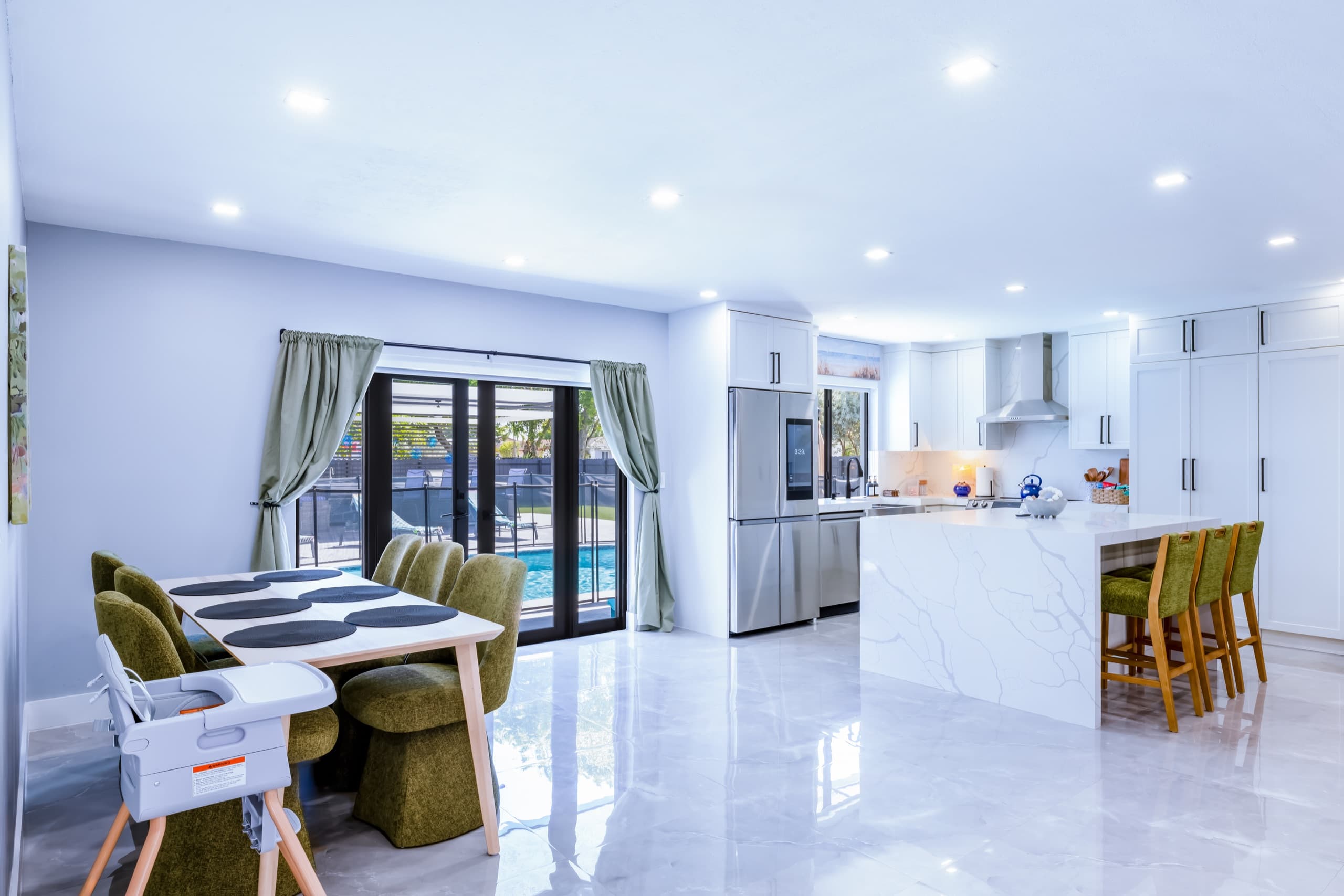 Dining table with green upholstered chairs and a high chair beside glass doors that look out to the pool, with the kitchen island at right.