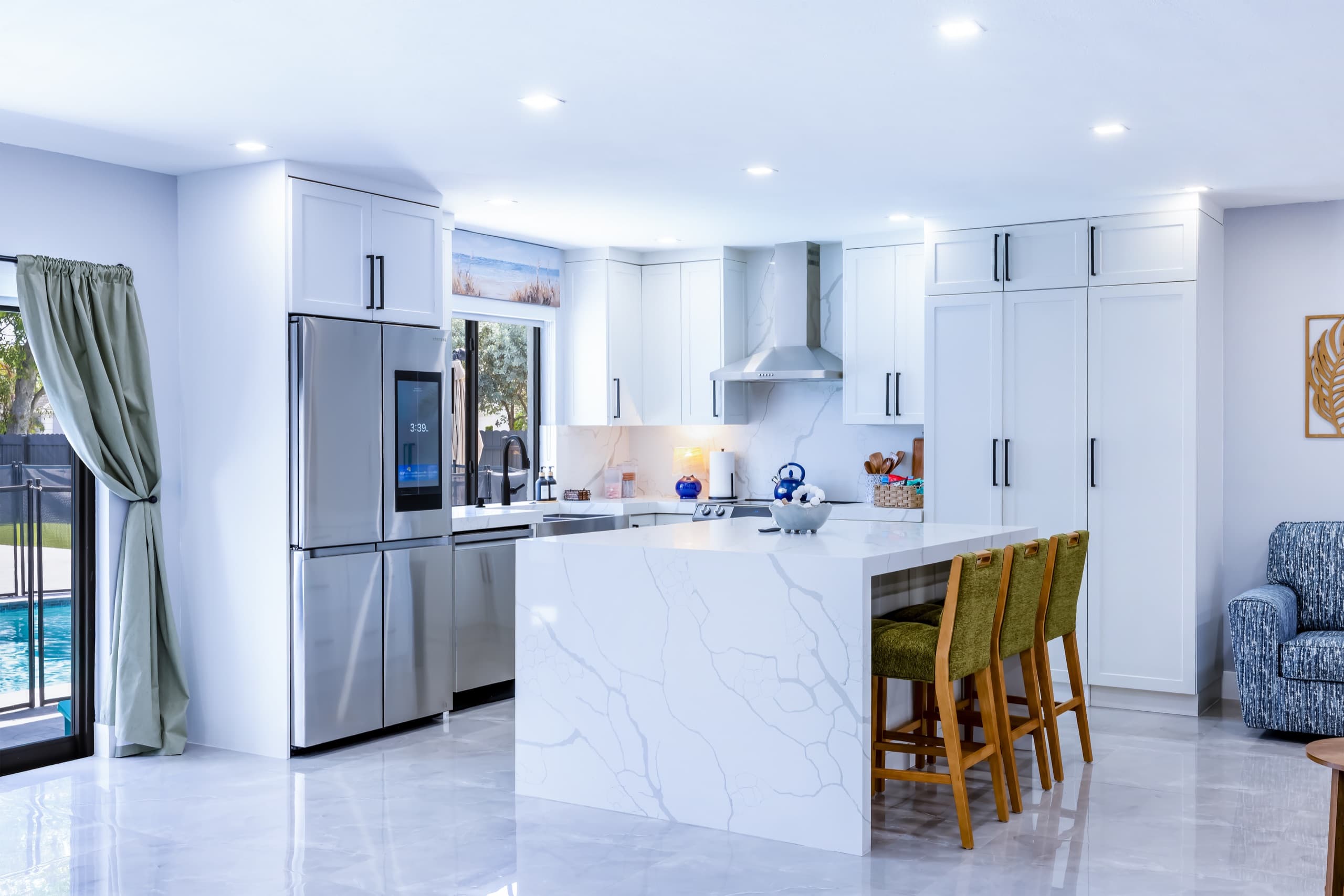 White kitchen with a veined white island, three green bar stools, a stainless refrigerator and a range hood.