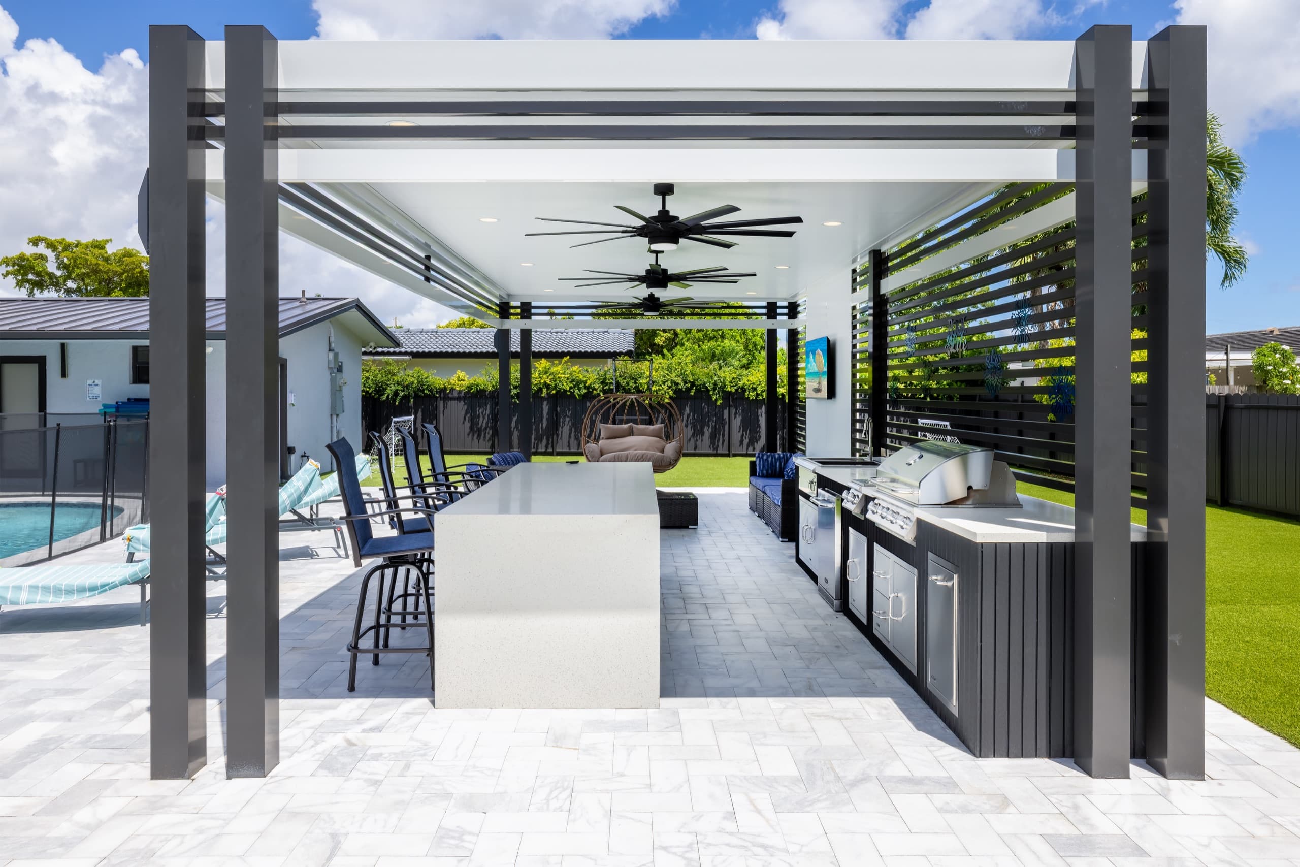 Outdoor kitchen pavilion seen head-on: a white roof on charcoal columns, a bar island with blue stools, a grill and three ceiling fans.