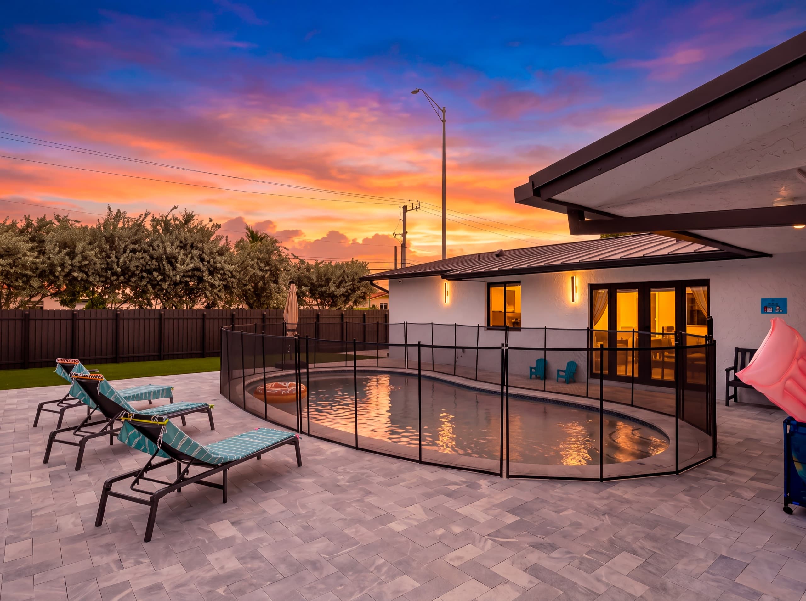 Loungers beside the fenced pool at dusk, with the lit house behind.