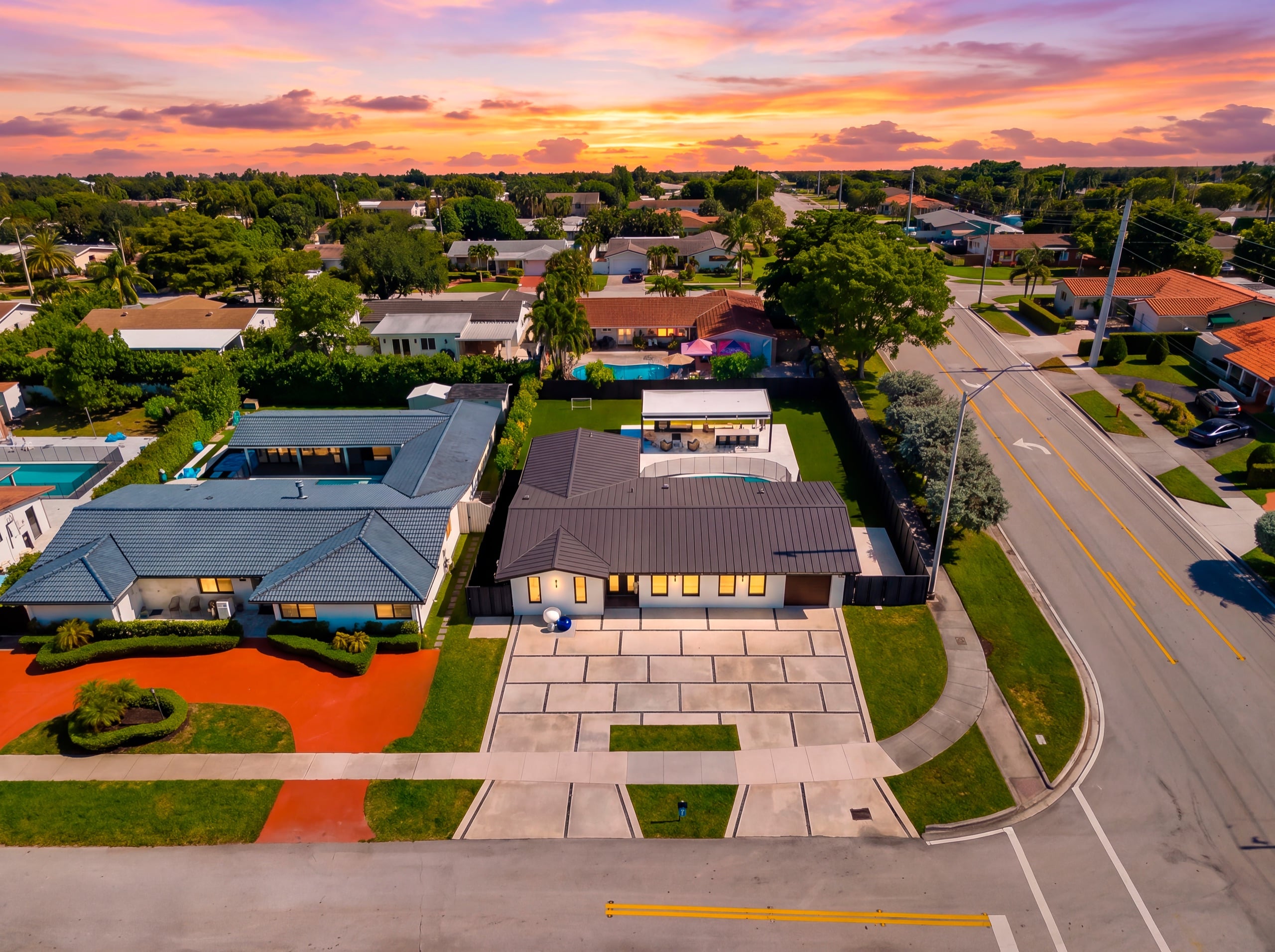 Aerial view of the front of the white single-storey house at dusk, with its windows lit and the pavilion and pool behind the roofline.