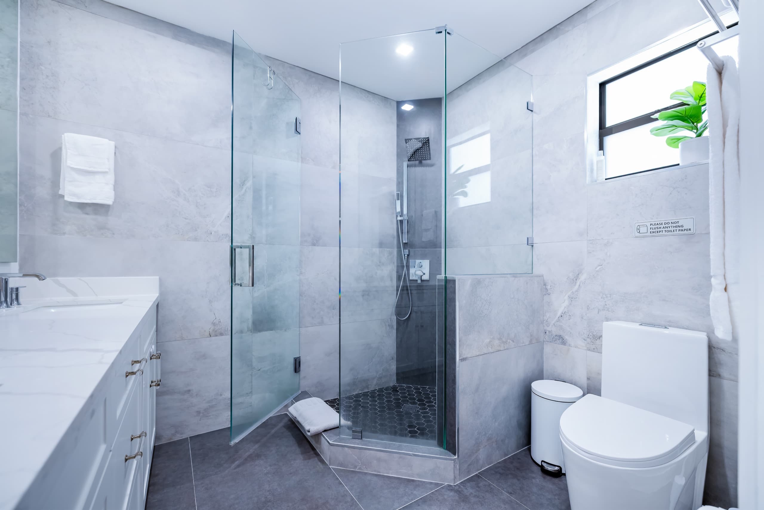 Bathroom with a frameless glass enclosure, grey stone-look tile and a window above the toilet.