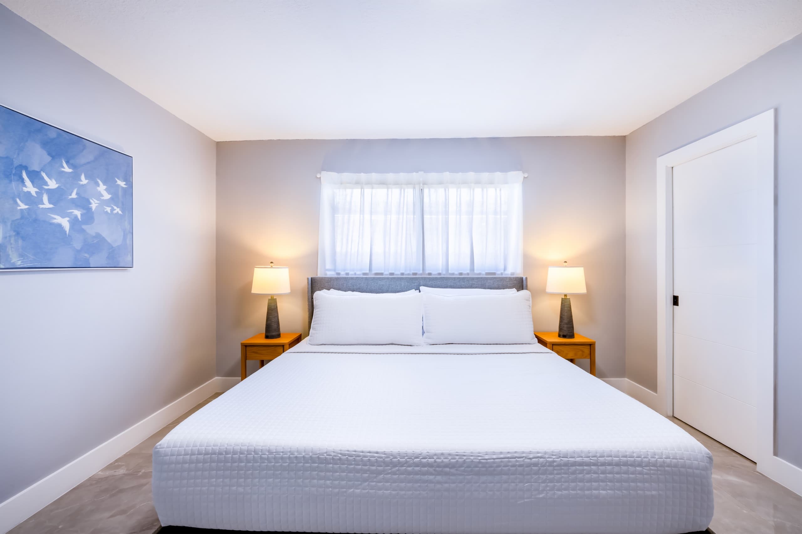 King bed with a grey upholstered headboard centred under a window between two lit lamps, with a blue bird painting at left.