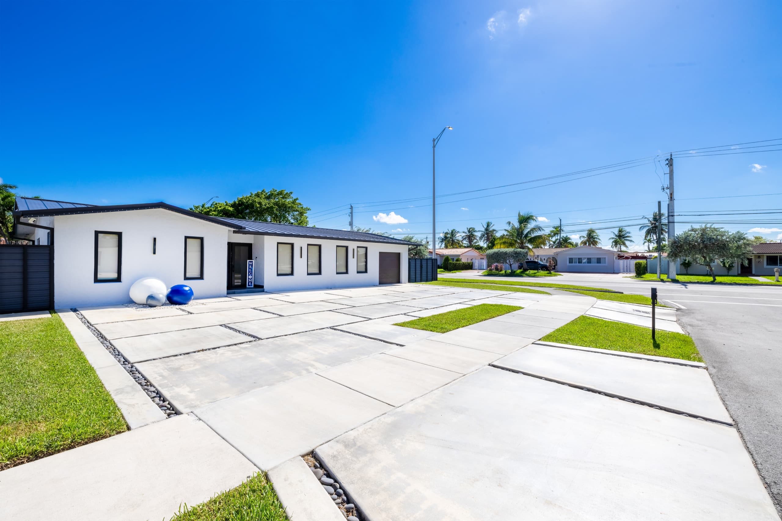 White single-storey house with a dark metal roof seen from the street corner, with a driveway of large concrete slabs leading to the front door.