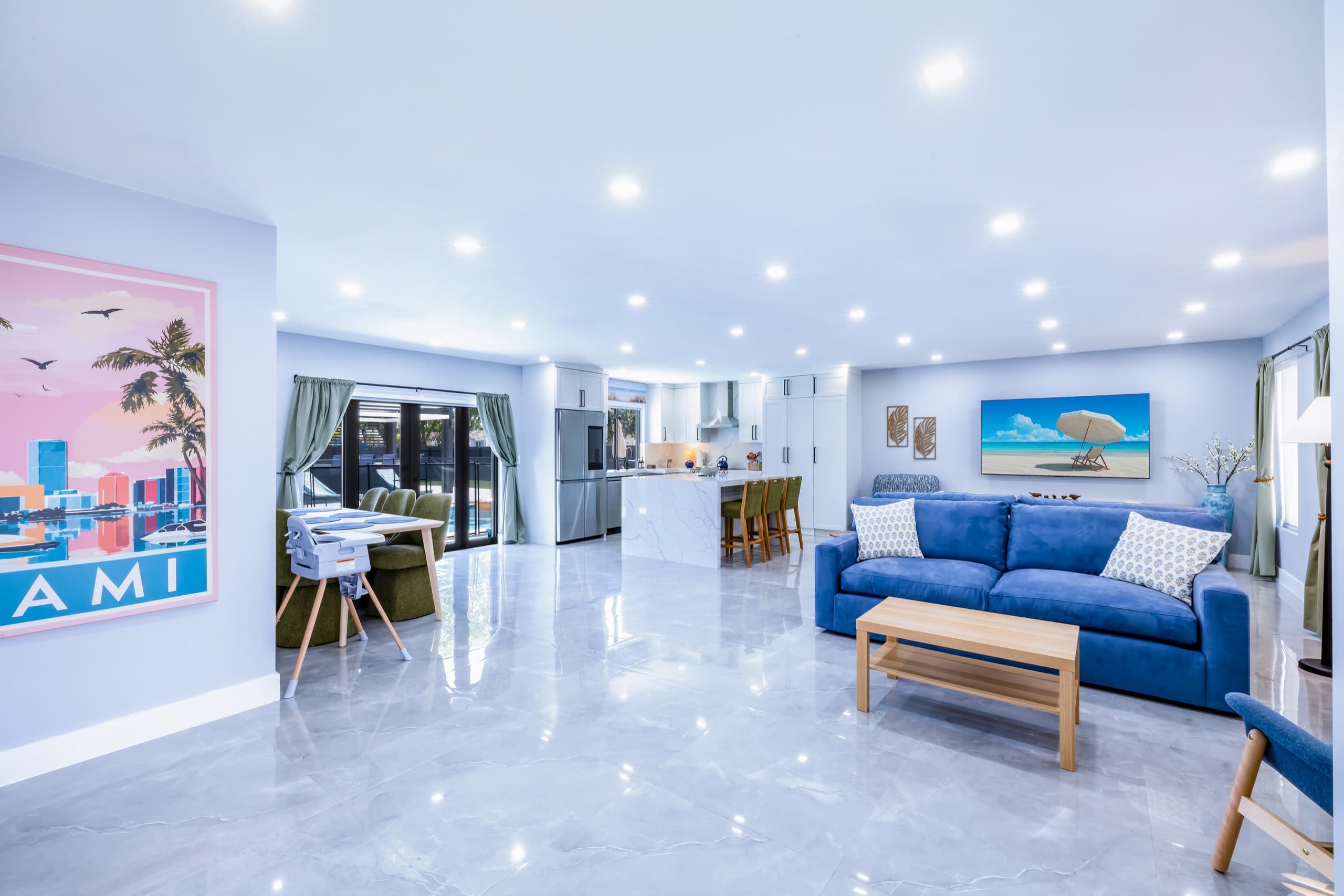 Open living, dining and kitchen area with a blue sofa and glossy grey floor, and glass doors looking out to the pool.