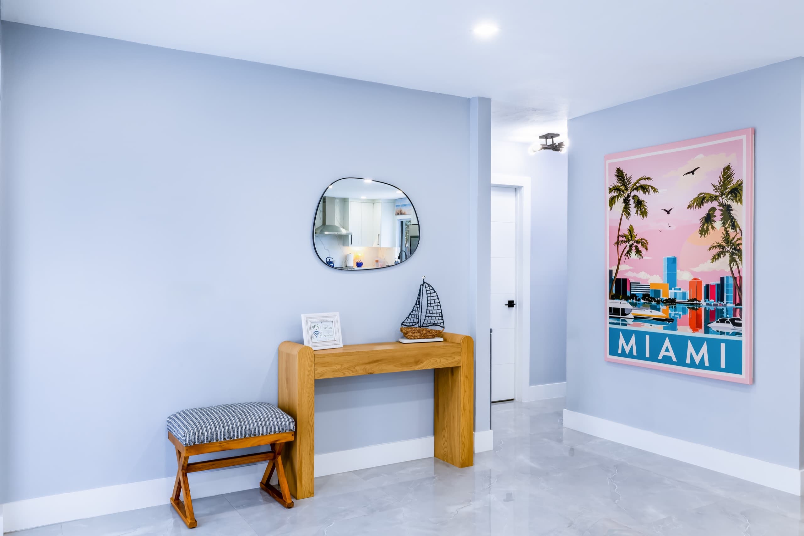 Entry area with a wooden console, a round mirror and a pink-and-blue Miami poster on a pale blue wall.